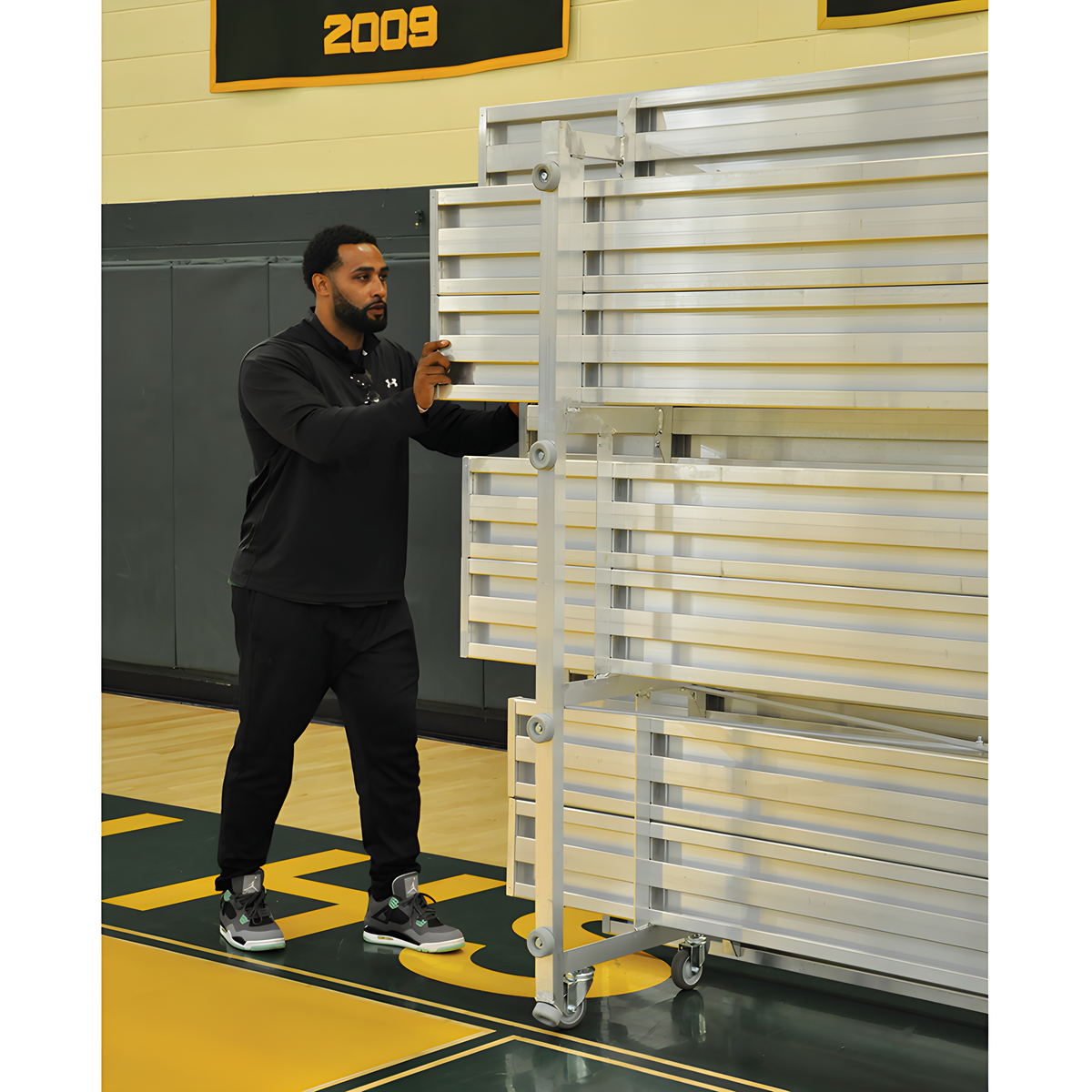 Man moving folded aluminum bleachers on wheels in school gymnasium.