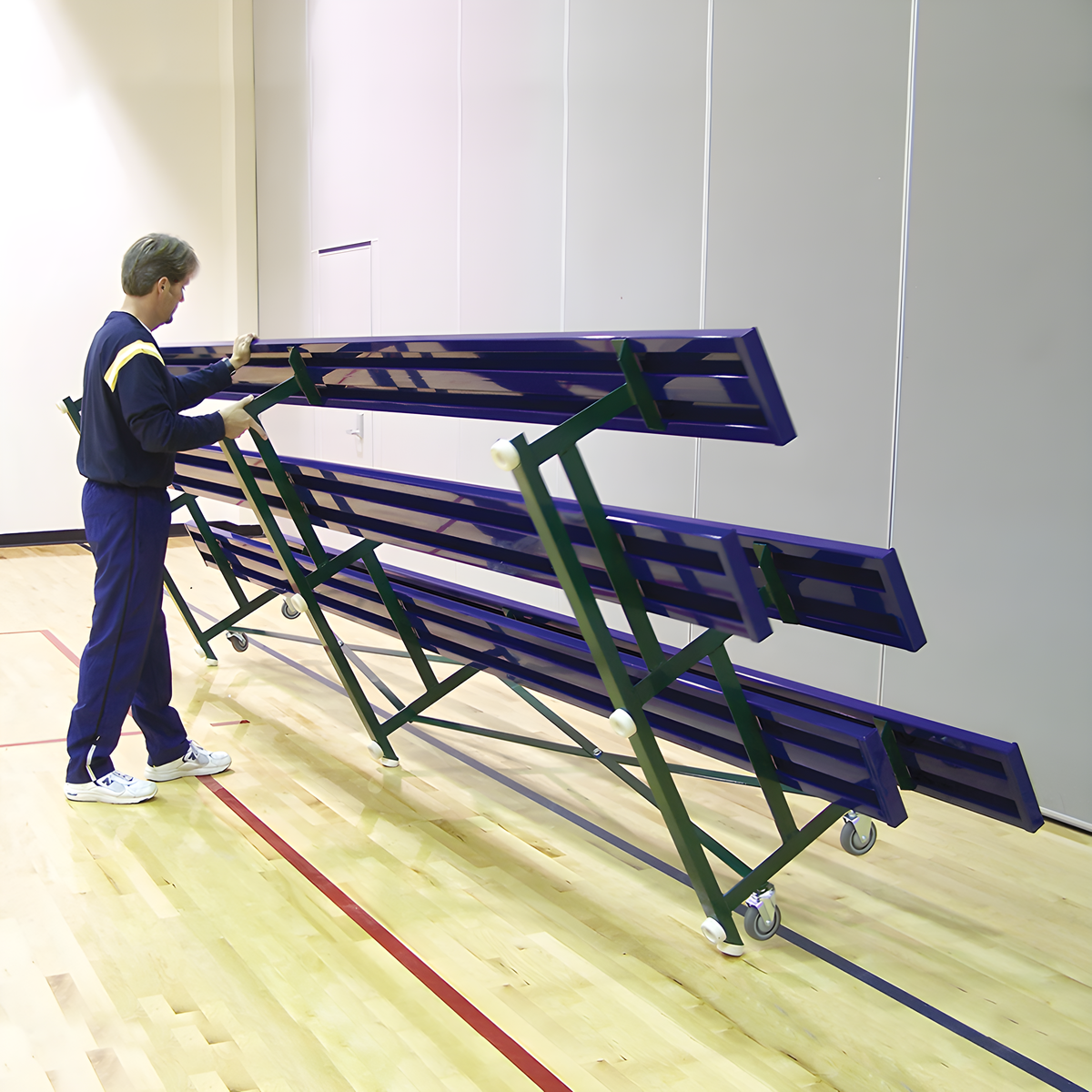 Man folding indoor bleacher seats with green frame and wheels.