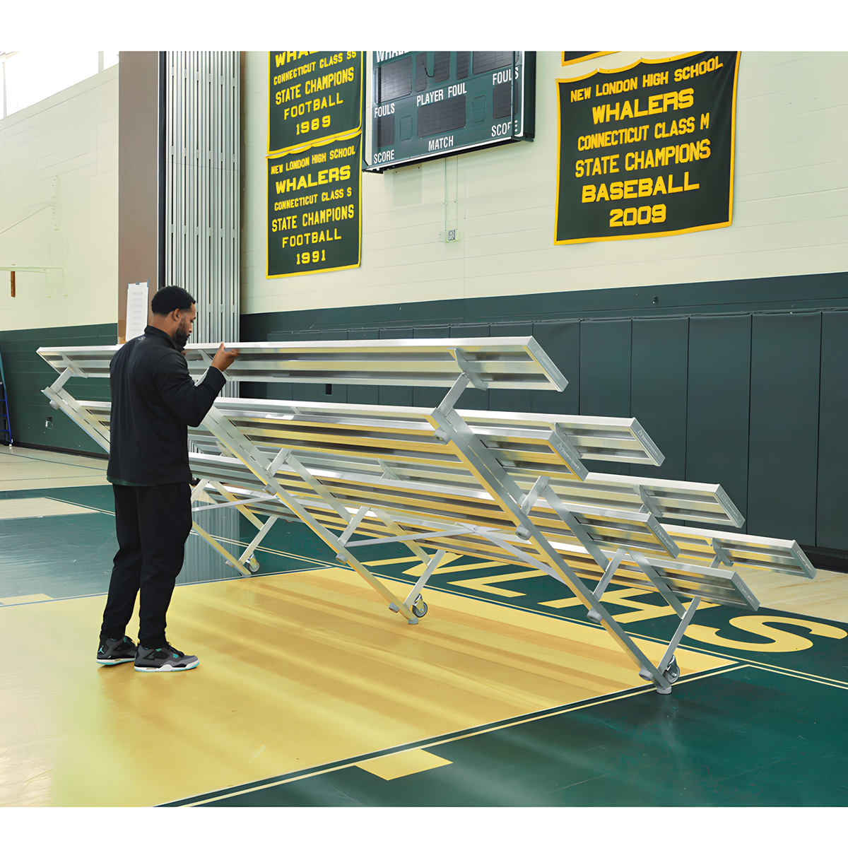 Man folding metal bleachers in gym with championship banners displayed.