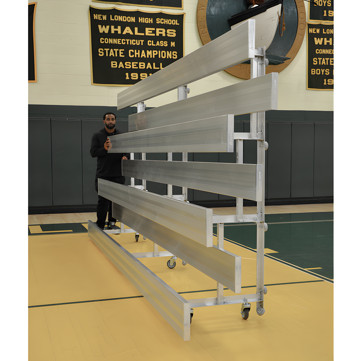 Man standing beside folded aluminum bleachers in gym with banners.