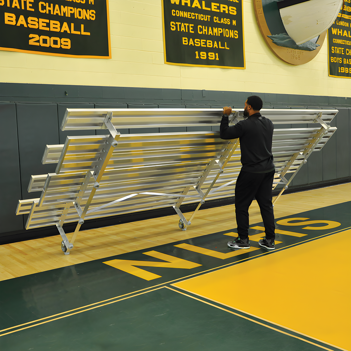 Man folding indoor bleacher seats with green frame and wheels.