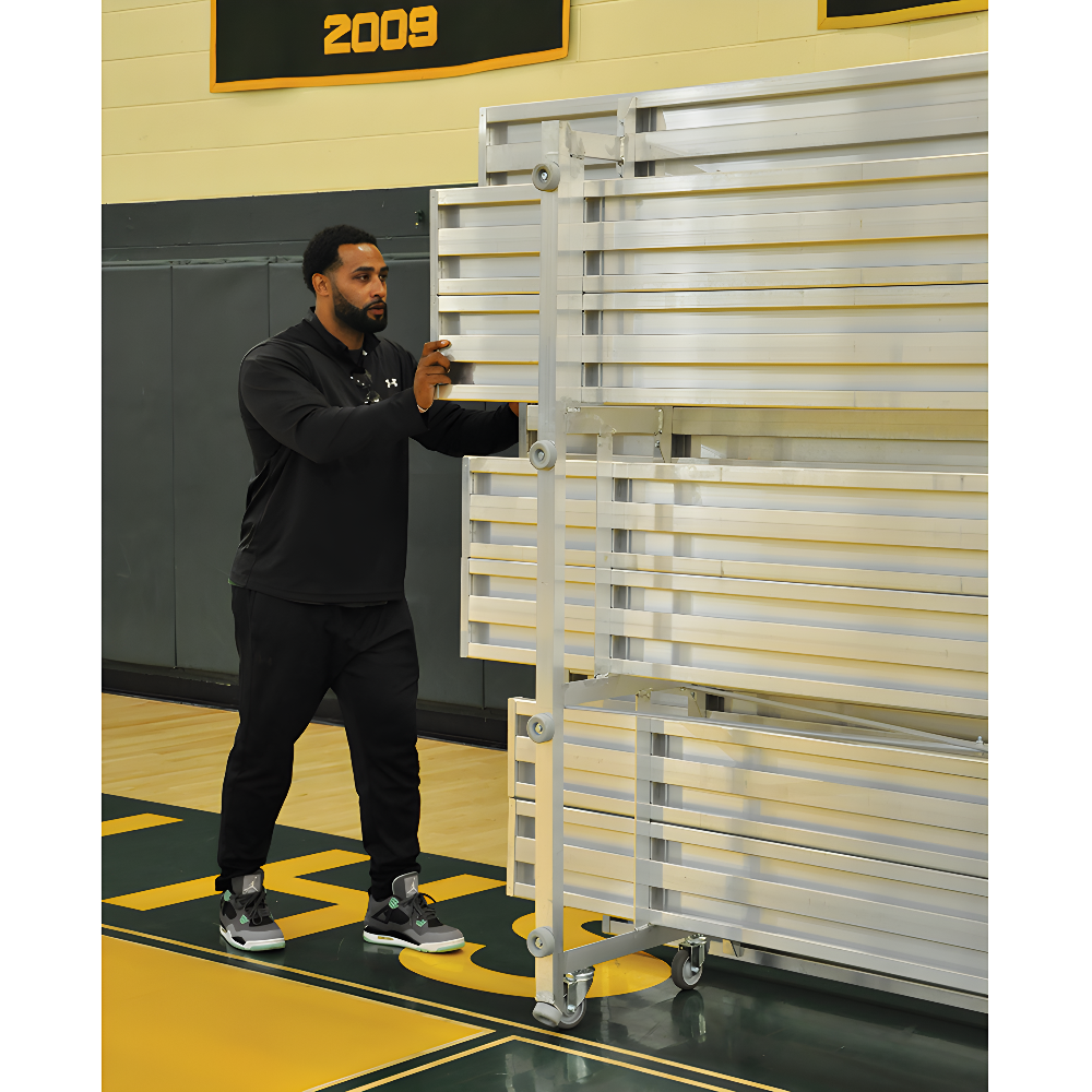 Man pushing folded indoor bleacher seats on wheeled aluminum frame.