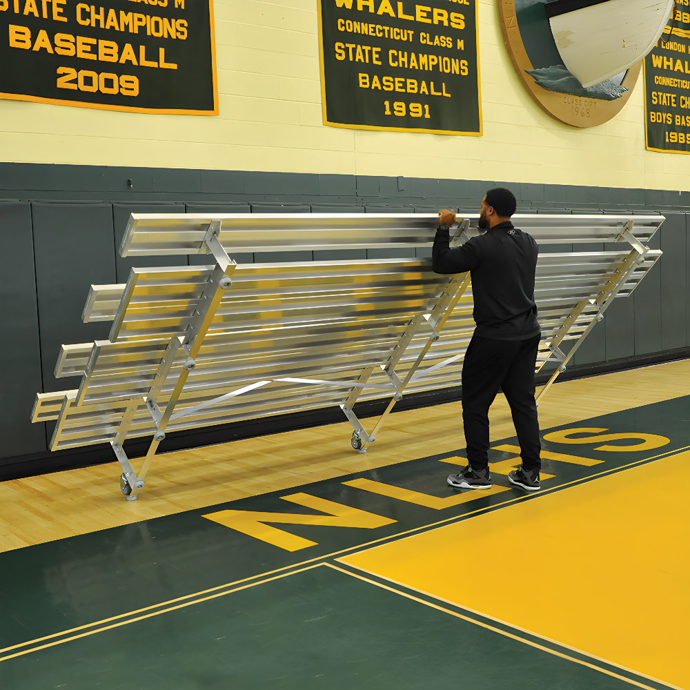 Man folding indoor bleacher seats in gym with championship banners.